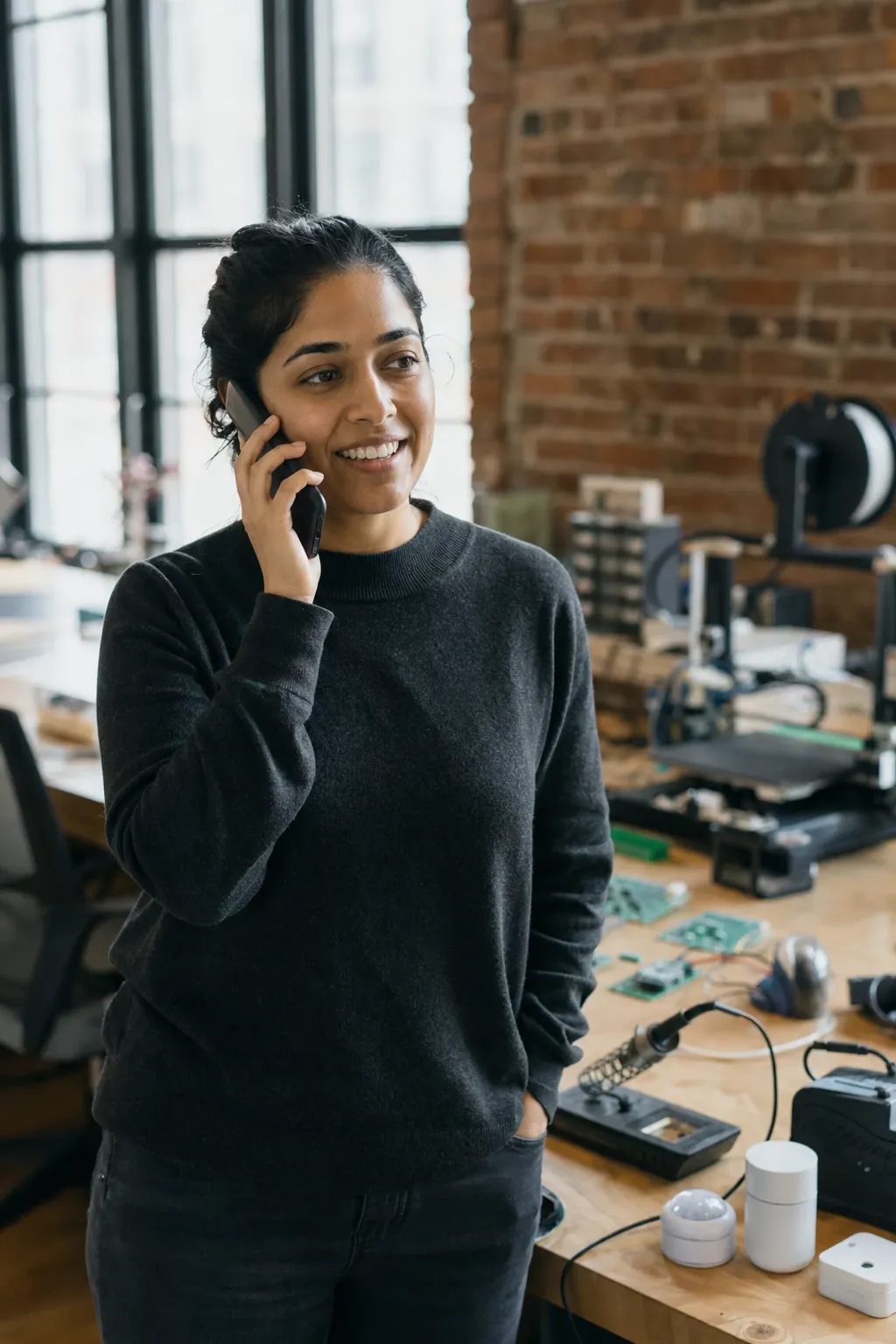 A startup founder on a phone call in a small hardware workshop, prototypes on the bench behind her